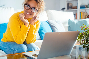 Attractive woman sitting on white sofa at home smiles, looks to her right in front of her laptop....
