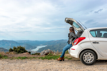 Woman traveler sitting on hatchback car with mountain background.