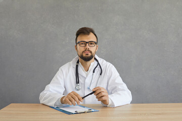 Portrait of hospital worker or doctor of general practice. Young man in white medical coat and glasses sitting at office desk looking at camera with serious face expression isolated on grey background