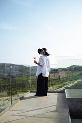Woman in elegant attire and a wide-brimmed hat enjoys a glass of rose wine while overlooking a...