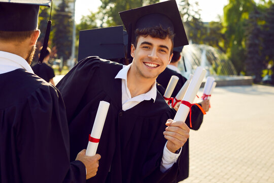 Portrait of happy smiling school, college or university student on graduation day. Young man in cap and gown with paper diploma scroll in hand looks back at camera while standing in line with others