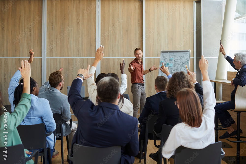 Wall mural Young colleagues raising hands to ask questions during business meeting in office. Diverse business people voting at the conference in meeting room listening their young male speaker.