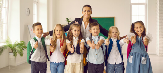 Positive primary school pupils boys and girls showing thumbs up posing together with woman teacher for educational promotion banner or kid additional training recommendation, stands in classroom