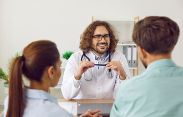 Obraz premium Cheerful friendly male doctor talking to patients about IVF treatment in his clinic. Happy smiling man in white medical coat and glasses sitting at table and looking at young family couple