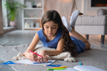 Beautiful caucasian preteen little girl daughter doing her homework task exercise drawing picture with felt-tip pen pencil while lying on floor white carpet at home. - obrazy, fototapety, plakaty