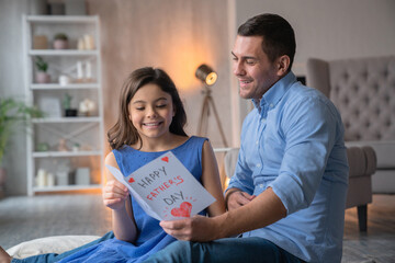 Happy Father's Day! Cute little preteen girl daughter greeting and gives handmade paper card for her attractive young father dad . Family sitting on the floor, cuddling and reading congratulations