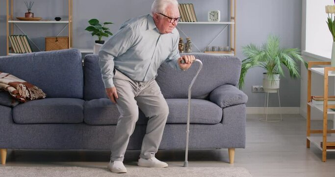 Portrait of senior gray-haired elderly injured man trying to get up from the couch with the help of his crutches having a back pain sitting on sofa in living room at home. 4k video.