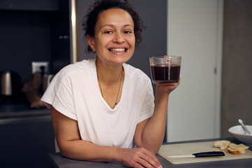 Happy smiling young woman in white pajama, standing at kitchen countertop and drinking coffee at home interior