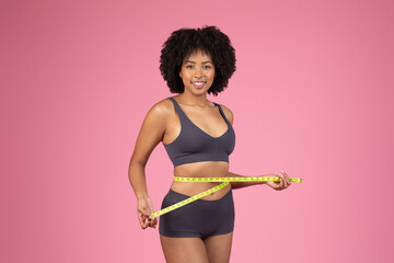 Smiling African American lady measuring waist on pink background