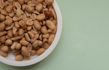 A bowl of peanuts on a green background. The peanuts are scattered all over the bowl and some are missing. Concept of abundance and variety