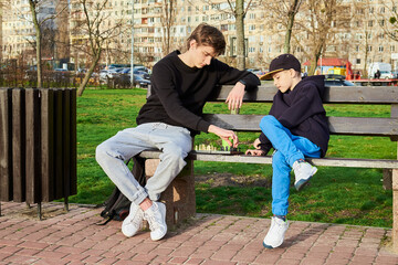 Teenager and an 8-year-old boy, are sitting on park bench playing chess. The teenager is teaching...
