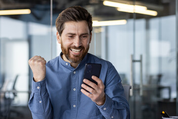 Happy and smiling young man sitting in office modern center and looking at phone, enjoying success and showing victory gesture with hand. Close-up photo