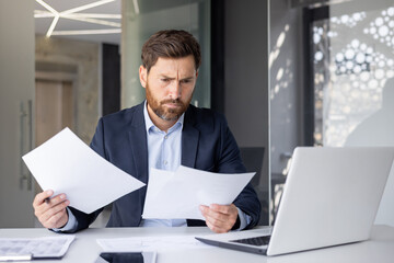 A confident young man businessman works in the office with a laptop and documents, sits at a desk...