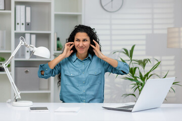 A professional woman in a blue shirt feeling overwhelmed by noise at her office desk with a laptop,...