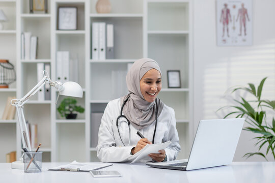 A cheerful Muslim female doctor wearing a hijab engages in an online consultation, taking notes and smiling in a well-organized medical office.
