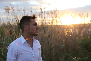A man in a white shirt stands in a field of tall grass. The sky is cloudy and the sun is setting