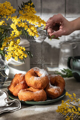 Delicious homemade crumpets or fried donuts, woman hand is sprinkling with powdered sugar on gray tile kitchen background in hard light