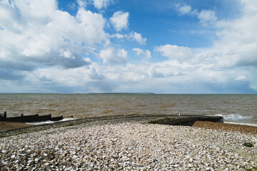 Oyster shells on Whitstable beach next to a slipway with a wooden groyne to the left. The clouds are dramatic. The isle of Sheppey can be seen on the horizon. - 785708921