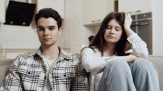 Young caucasian man sitting on a couch at home next to offended young caucasian woman and looking at the camera. Concept of problems in couple relationship