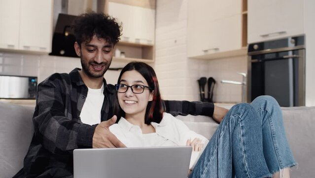 Young man in casual clothes sitting on a couch in a kitchen and showing something with passion on a laptop to his smiling girlfriend. Concept of happiness and harmony in couple relationship