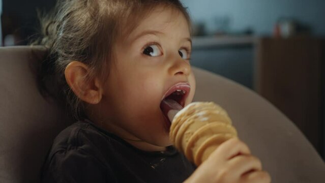 Pretty little girl eating an ice-cream indoors, at home sitting on high-chair. Cute kid with tasty sweet food. Baby girl enjoying ice cream.