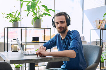 Portrait of young handsome man with headphones working with laptop, looking at camera