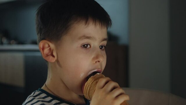 Close up view of a little boy eating Ice cream in cone sitting at home. Small child eating plombir and cream messy on her mouth. Cute kid with tasty sweet food.