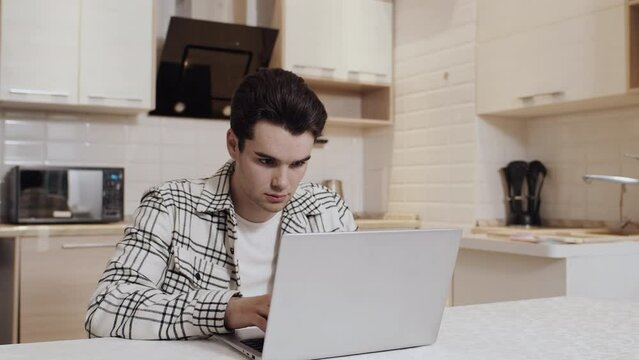 Young caucasian man in casual clothes working on a laptop in a kitchen when his girlfriend hugs him from the back. Concept of happiness and support in the couple relationship