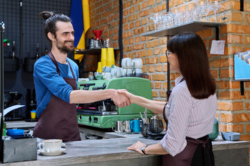 Young man in apron shaking hands with woman owner of coffee shop