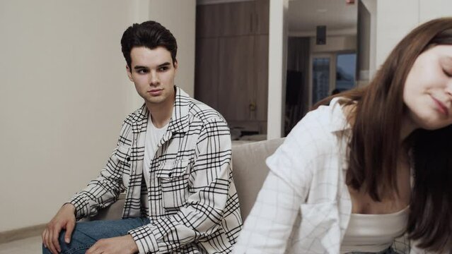 Offended young caucasian woman sitting on the couch back to young caucasian man who trying to apologize, but she refuses it. Concept of problems in couple relationship