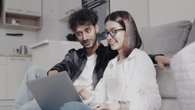Young smiling couple in casual clothes sitting on a couch in a kitchen and emotionally discussing something with a laptop pointing a finger in a screen. Concept of happiness and consent in the