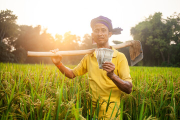 rural farm worker showing indian paper currency and holding a garden hoe on his shoulder standing...