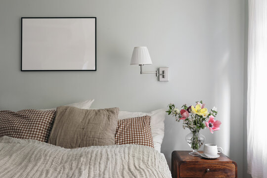 Elegant bedroom. Tulips, cherry tree blossoms bouquet in glass vase. Wooden night stand. Cup of coffee. Blank black picture frame mockup. Lamp, pleated shade on mint wall. Checkered pillows.