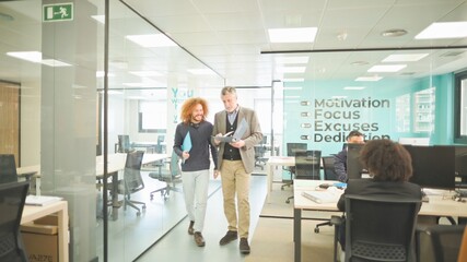Two professionals walking and discussing work in a dynamic office setting.