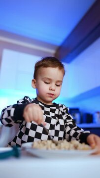 Cute Caucasian toddler sitting at high chair eats porridge. Baby takes a spoon to his mouth and tilts his head. Low angle view. Vertical video.