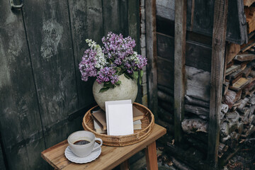 Moody rustic spring outdoor still life. Purple, white lilac flowers bouquet, textured vase. Cup of coffee, books. Greeting card, invitation mockup. Blurred old wooden door background. Pile of firewood