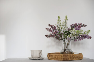 Moment of tranquility with cup of coffee, wicker tray. Floral bouquet of blooming purple and white lilacs branches,glass vase. Springtime breakfast scene, table. White wall background, selective focus