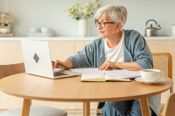 Online education courses webinar concept. Middle aged senior woman using laptop computer writing notes. Focused mature old woman enjoying studying online from home written records doing online work