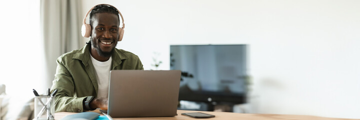 Positive black man in wireless headphones working online on laptop