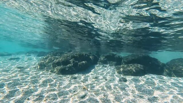 Dive towards underwater rock formations and fish swimming in crystal clear, pure blue water with light reflections on the seabed. Natural texture, high angle view. Underwater shot in 4K resolution.