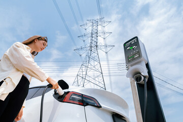 Woman recharge EV electric car battery at charging station connected to electrical power grid tower...