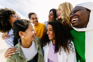 Seven happy young adult women from different cultures laughing together outdoors. Female friendship concept with diverse group of girls friends hugging each other having fun at city street