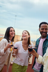 Vertical. Cheerful group multiracial young friends laughing holding wine glass at summer rooftop...