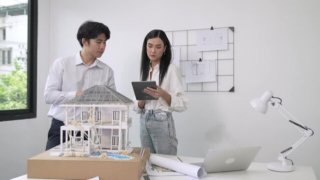 Beautiful young caucasian project manager brief asian engineer by using tablet while engineer measuring house model on meeting table with blueprint, architectural equipment scatter around. Immaculate