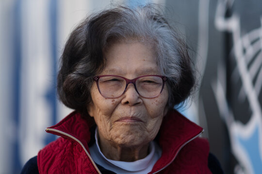 A ninety three year old woman with a splash of gray hair looks at the camera with a coy smile. She is Asian, of Okinawan (Ryukyuan) descent. She wears a red vest and is seated outdoors.
