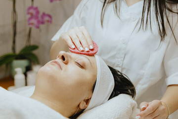 A girl cosmetologist cleanses the patient s face with a scrubbing sponge.