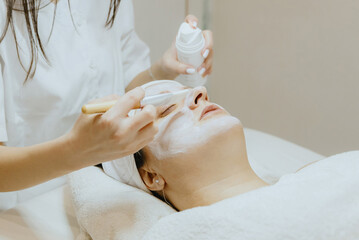 A girl cosmetologist applies a cream mask with a brush to a woman s face.