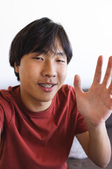 Asian teenage boy waving during a video call at home, sitting indoors