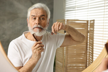 Senior man trimming mustache with scissors near mirror in bathroom