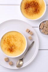 Delicious creme brulee in bowls, sugar cubes and spoon on white wooden table, flat lay